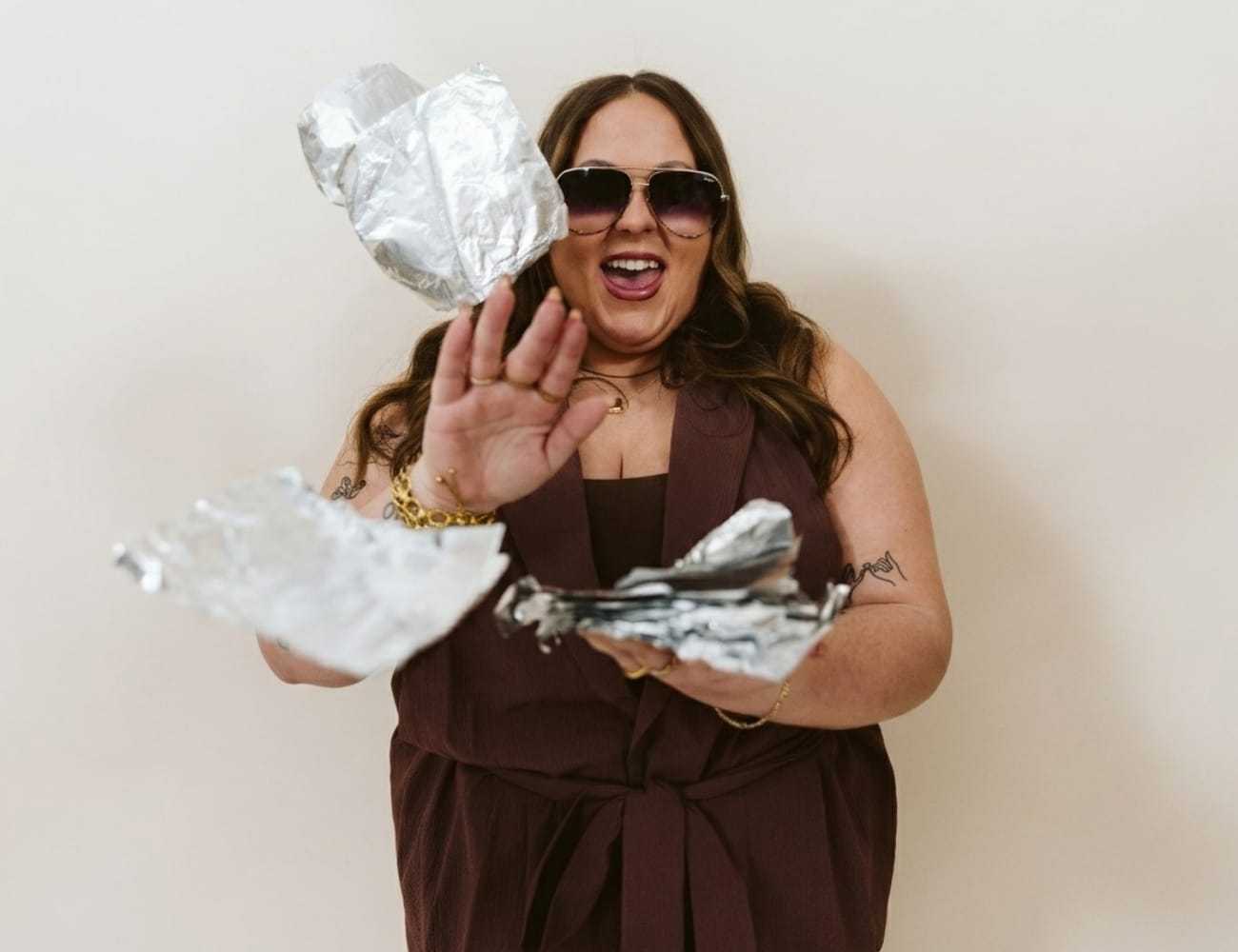 Woman joyfully tossing foil sheets in the air, wearing stylish sunglasses.